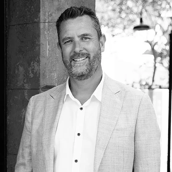 Smiling middle-aged man with short hair and beard wearing a light blazer and white shirt standing outdoors near a stone wall.