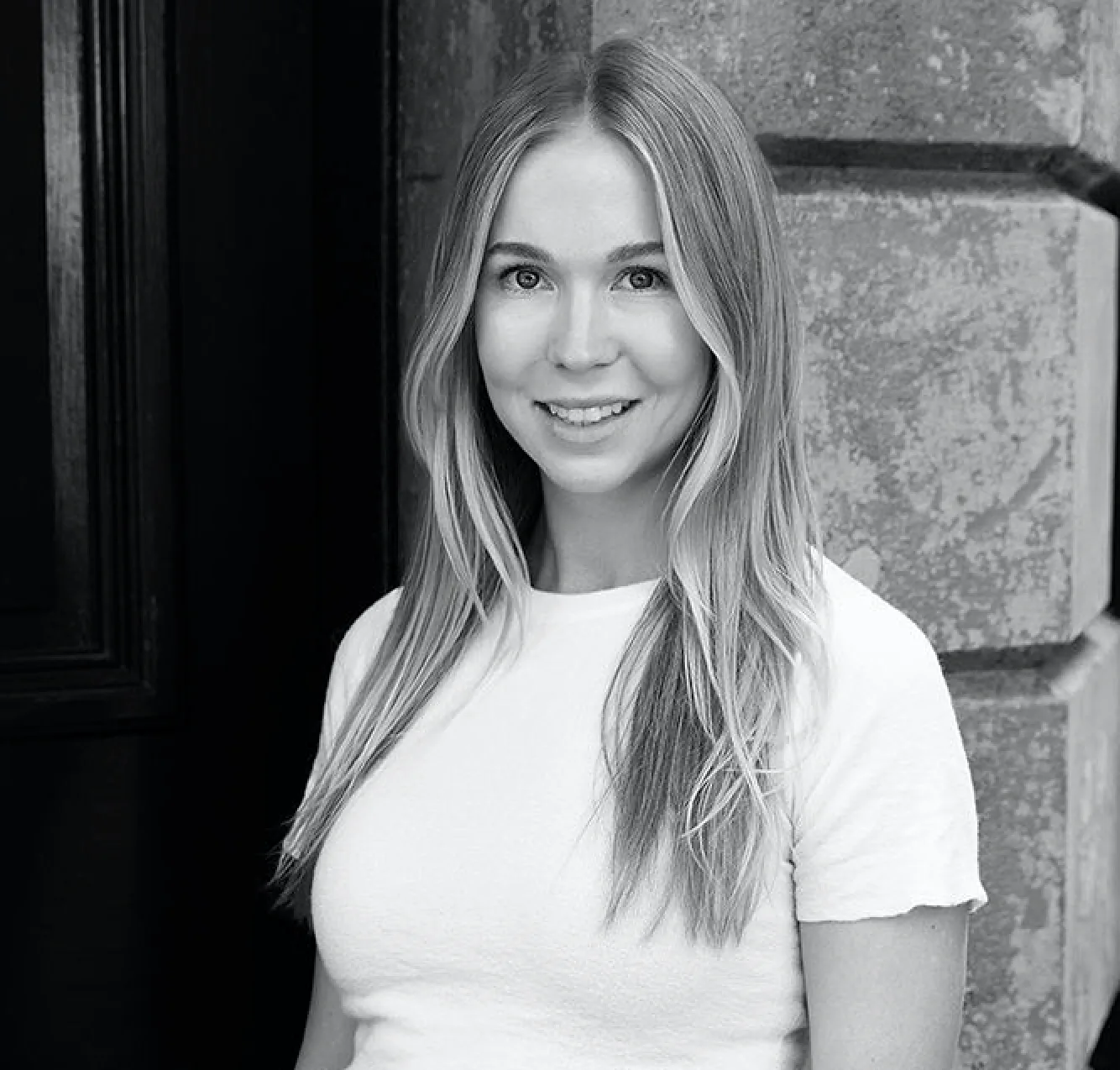 Smiling young woman with long blonde hair wearing a white shirt standing against a textured wall.