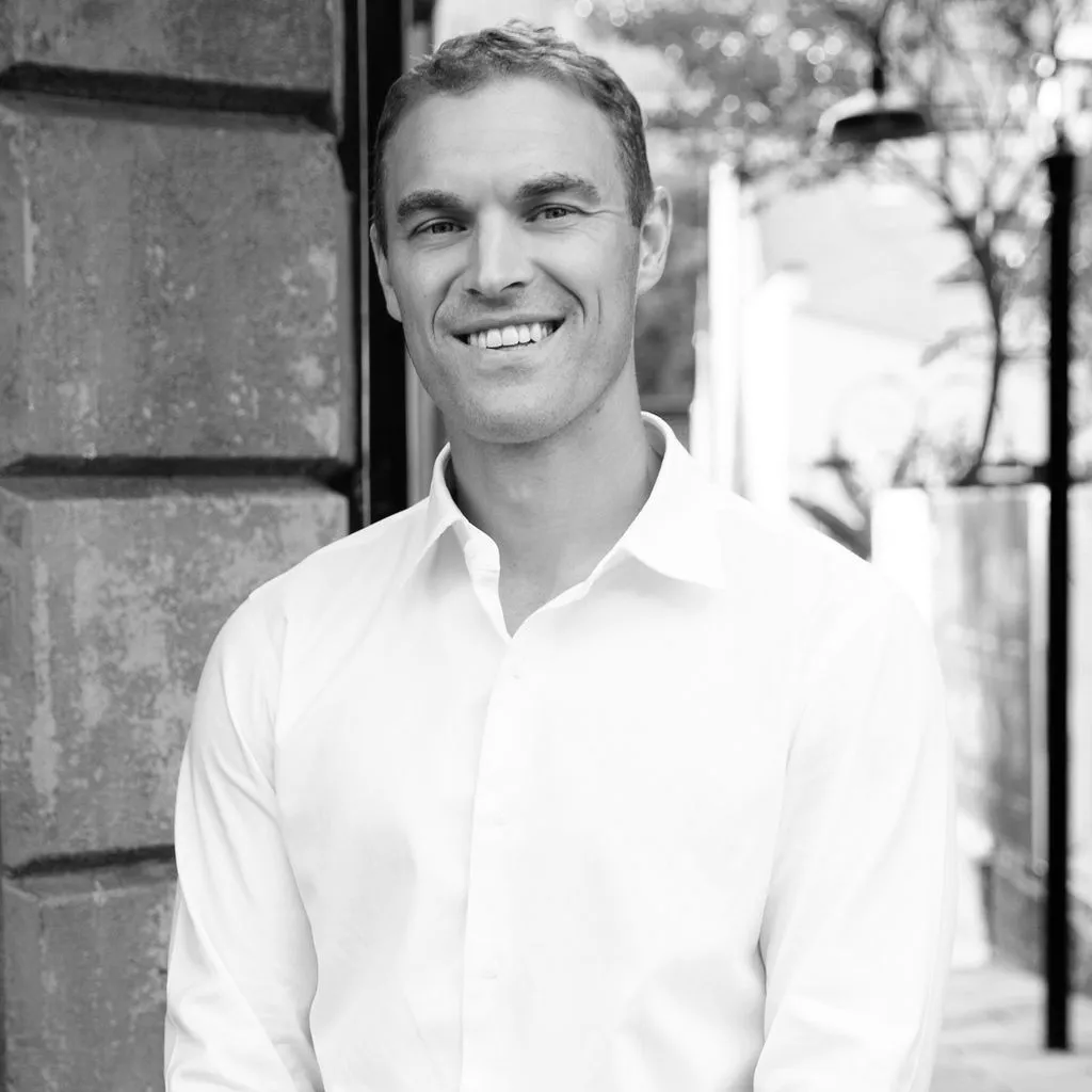 Smiling man in a white button-up shirt standing outdoors next to a textured wall.