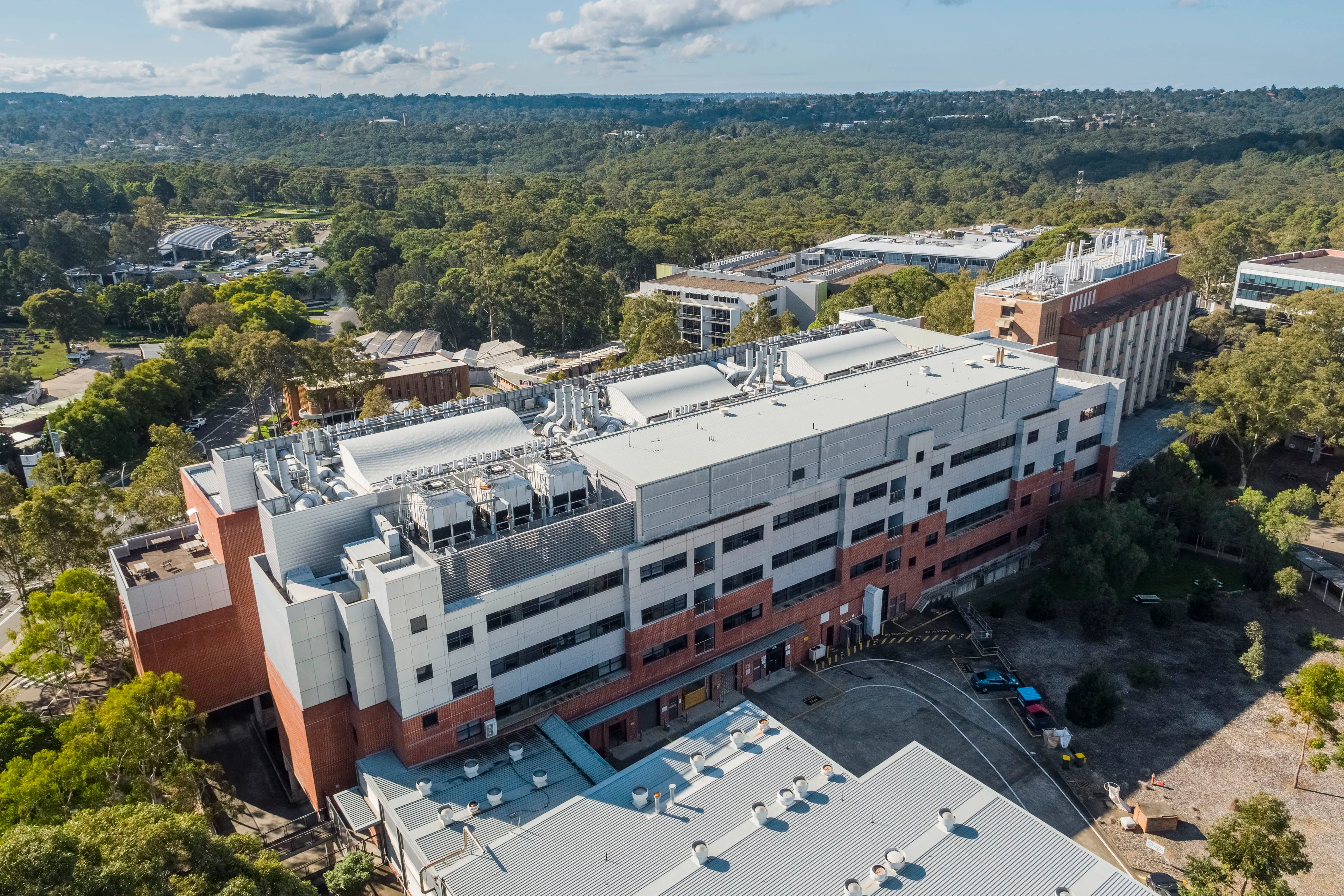 Aerial view of a large, multi-story research or industrial building with visible rooftop ventilation systems, surrounded by trees and greenery.