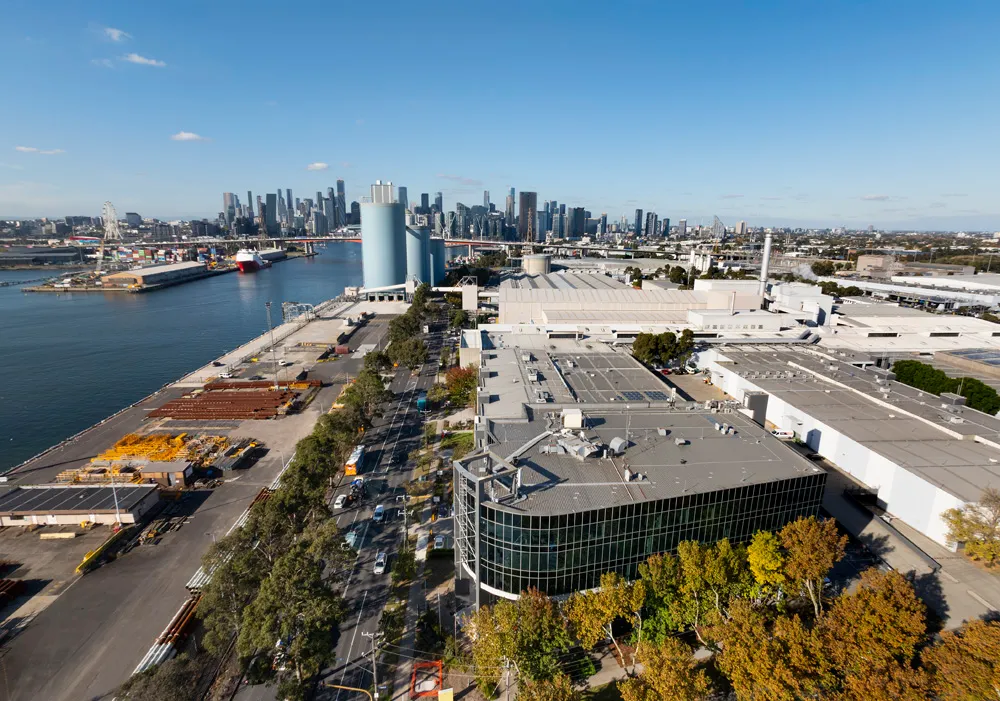 Aerial view of an industrial port area with warehouses, a road lined with trees, and a city skyline in the background under a clear blue sky.