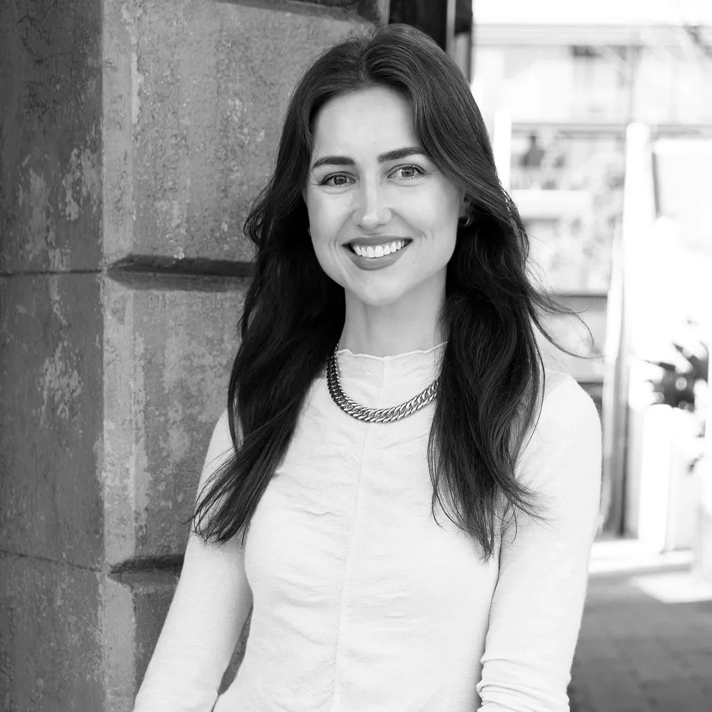 Smiling woman with long hair wearing a light-colored top and a chain necklace standing against a textured wall.