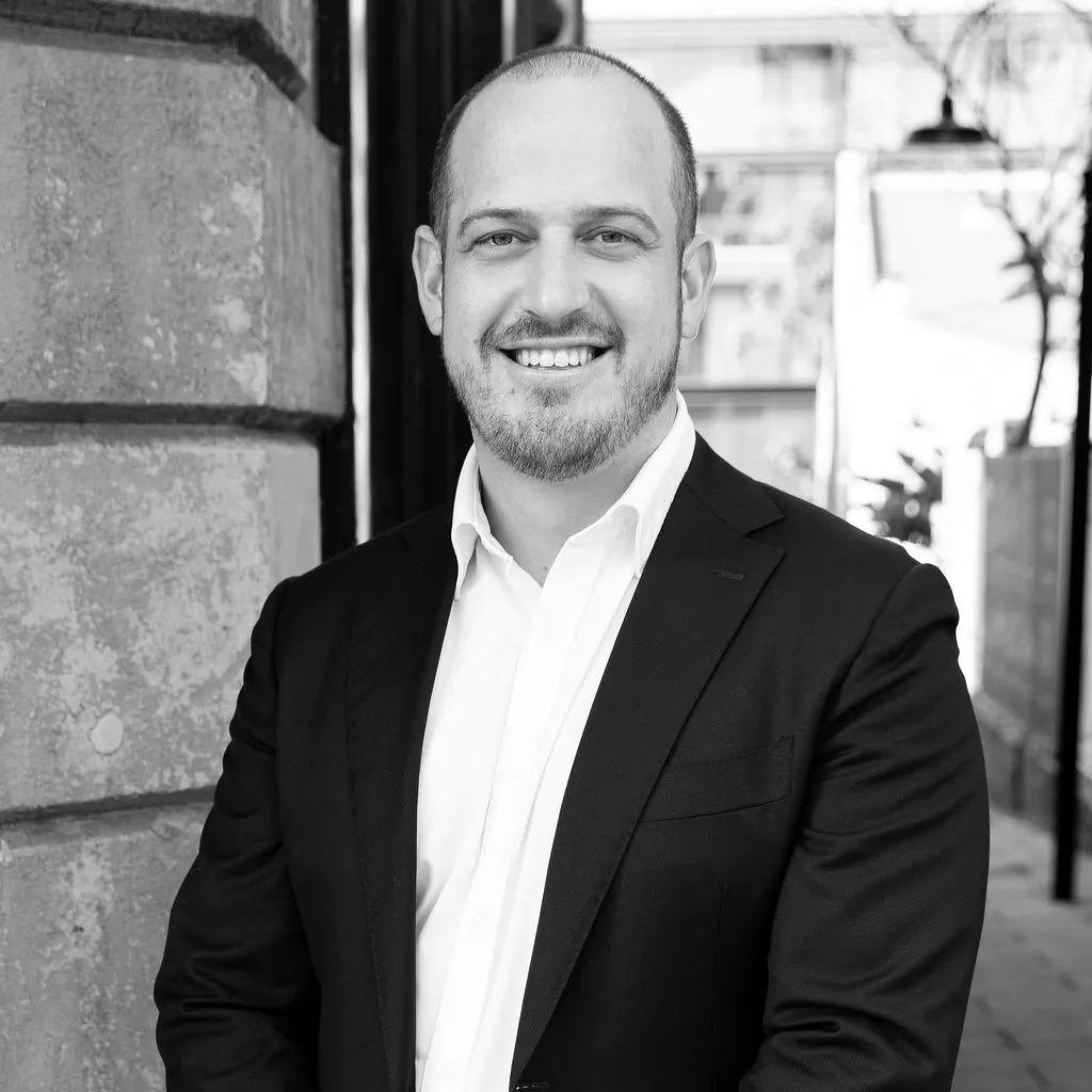 Smiling man in a white shirt and dark blazer standing outdoors near a textured stone wall.