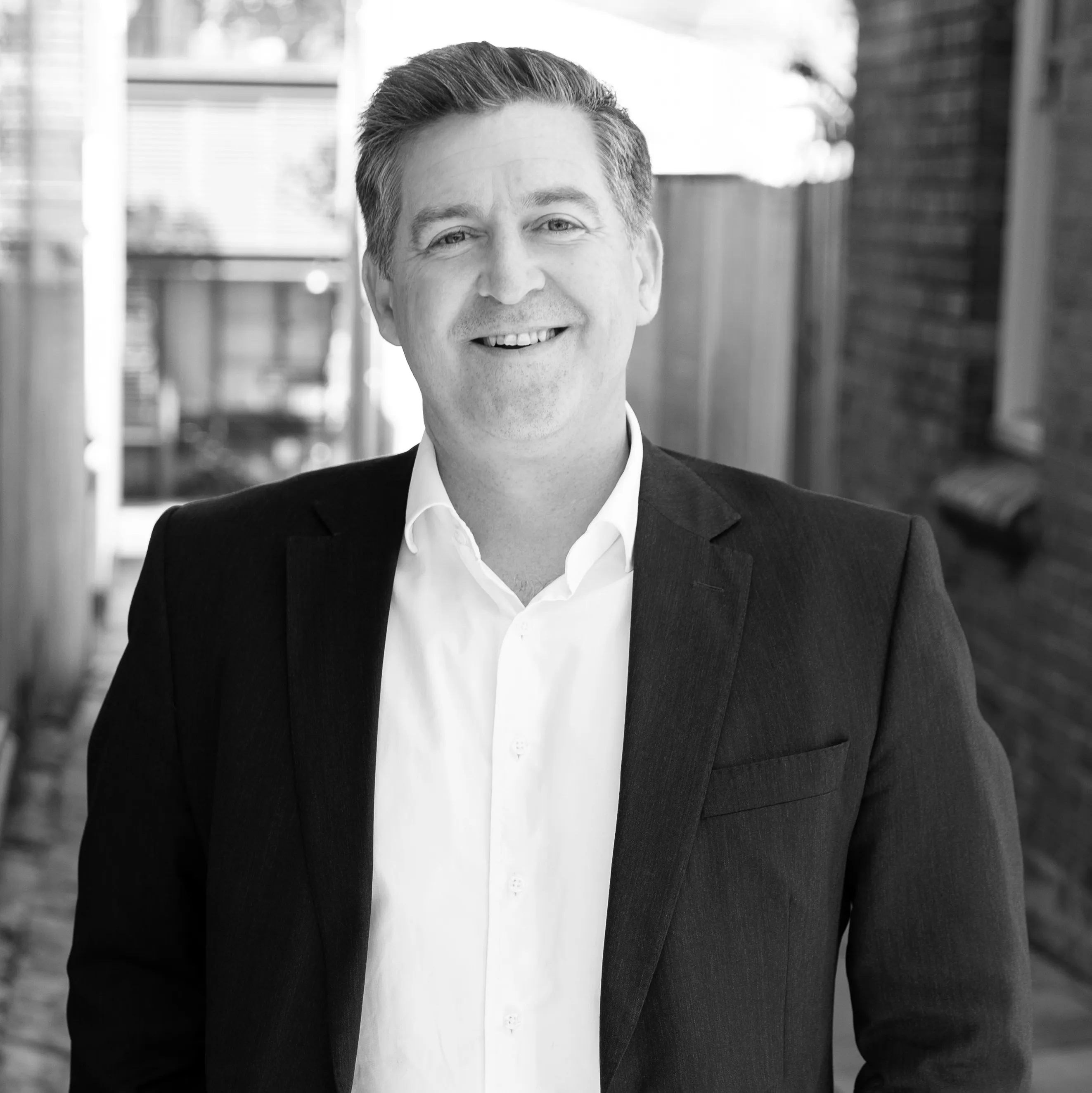 Smiling man wearing a dark suit jacket and white shirt standing outdoors in a narrow alleyway.