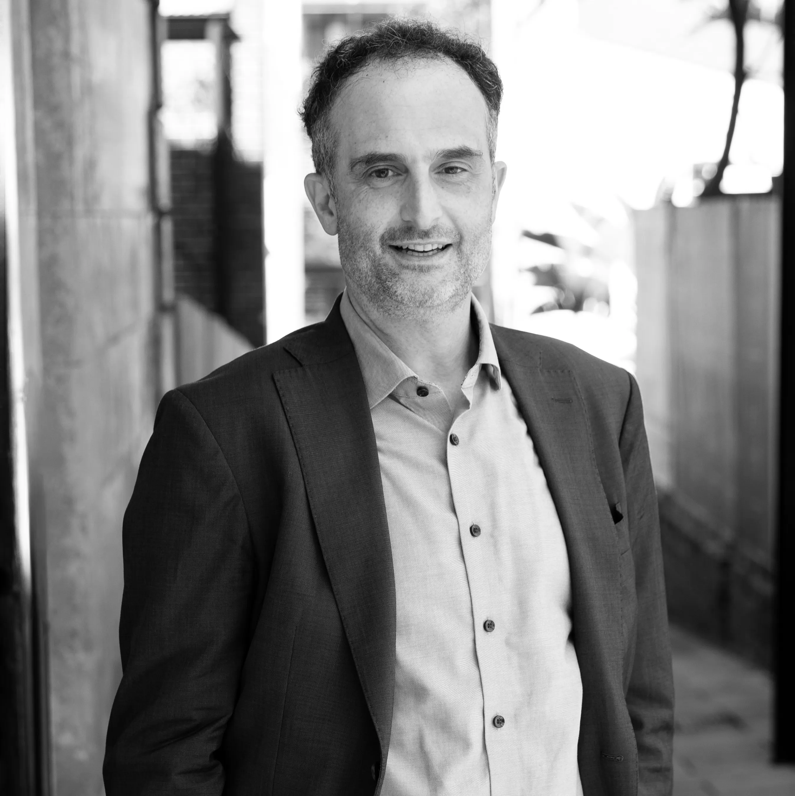 Smiling man with short curly hair wearing a suit jacket and button-up shirt in an urban outdoor setting.