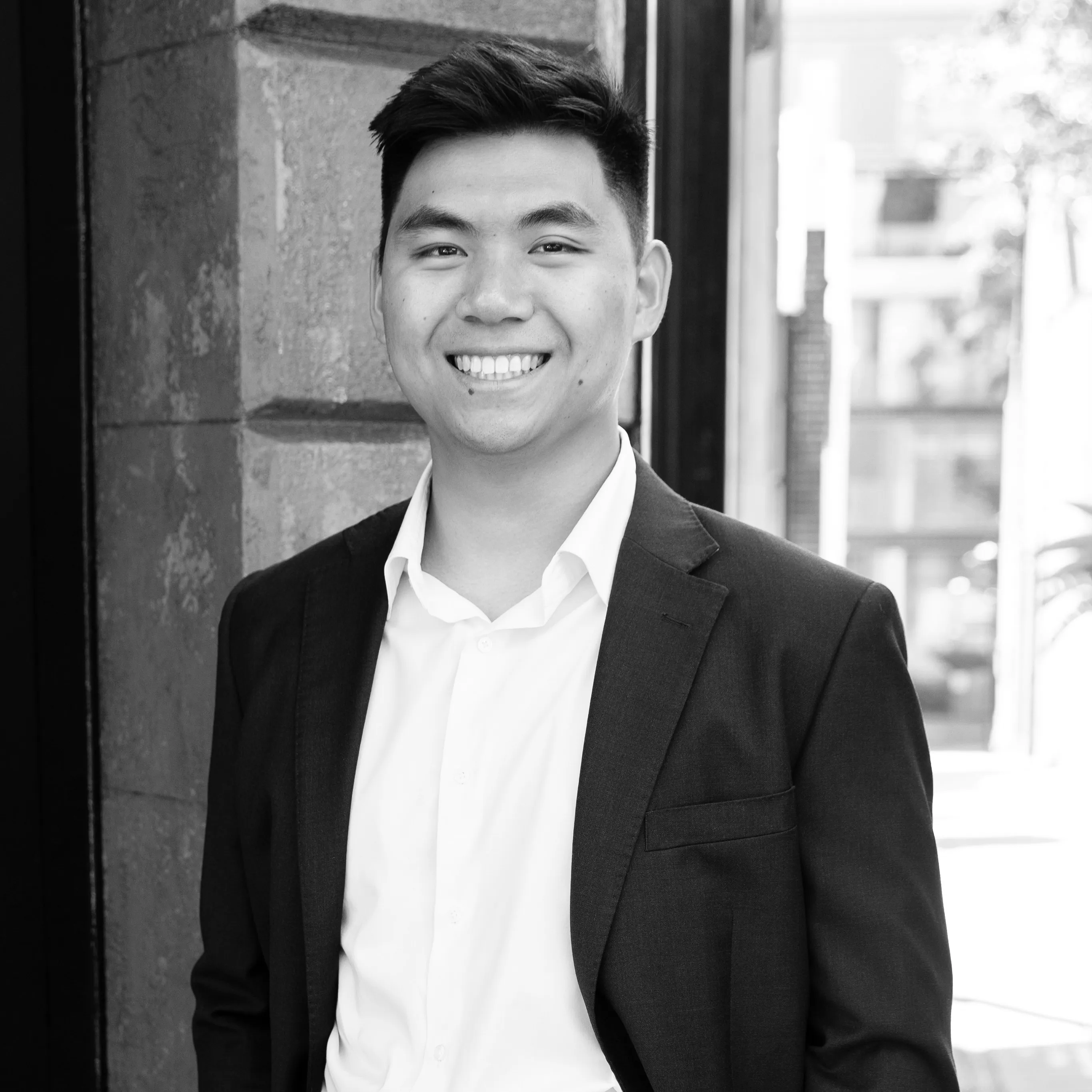 Smiling young man wearing a suit jacket and white shirt standing outdoors by a stone wall.