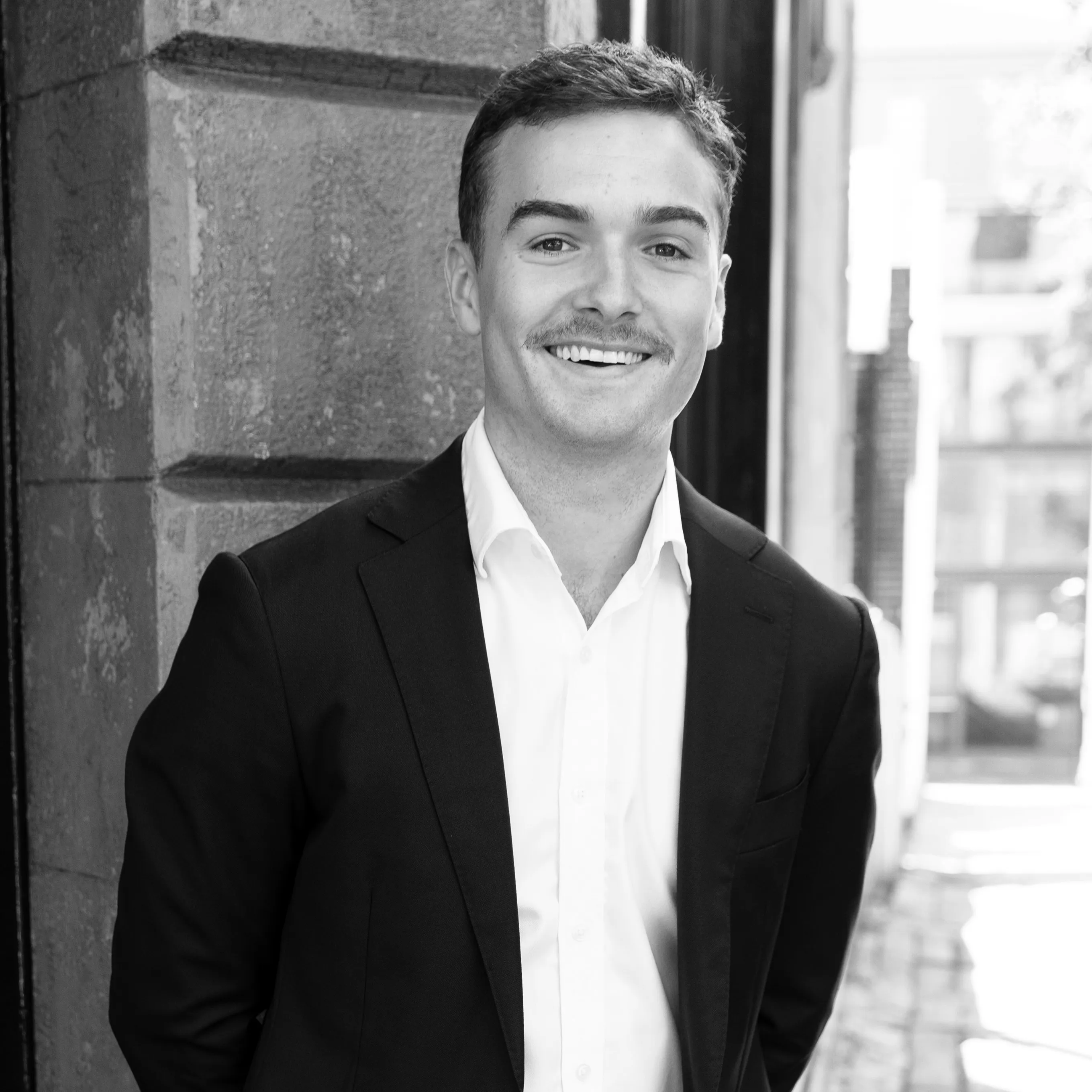 Smiling young man with a mustache wearing a dark suit and white shirt, standing outdoors near a textured stone wall.