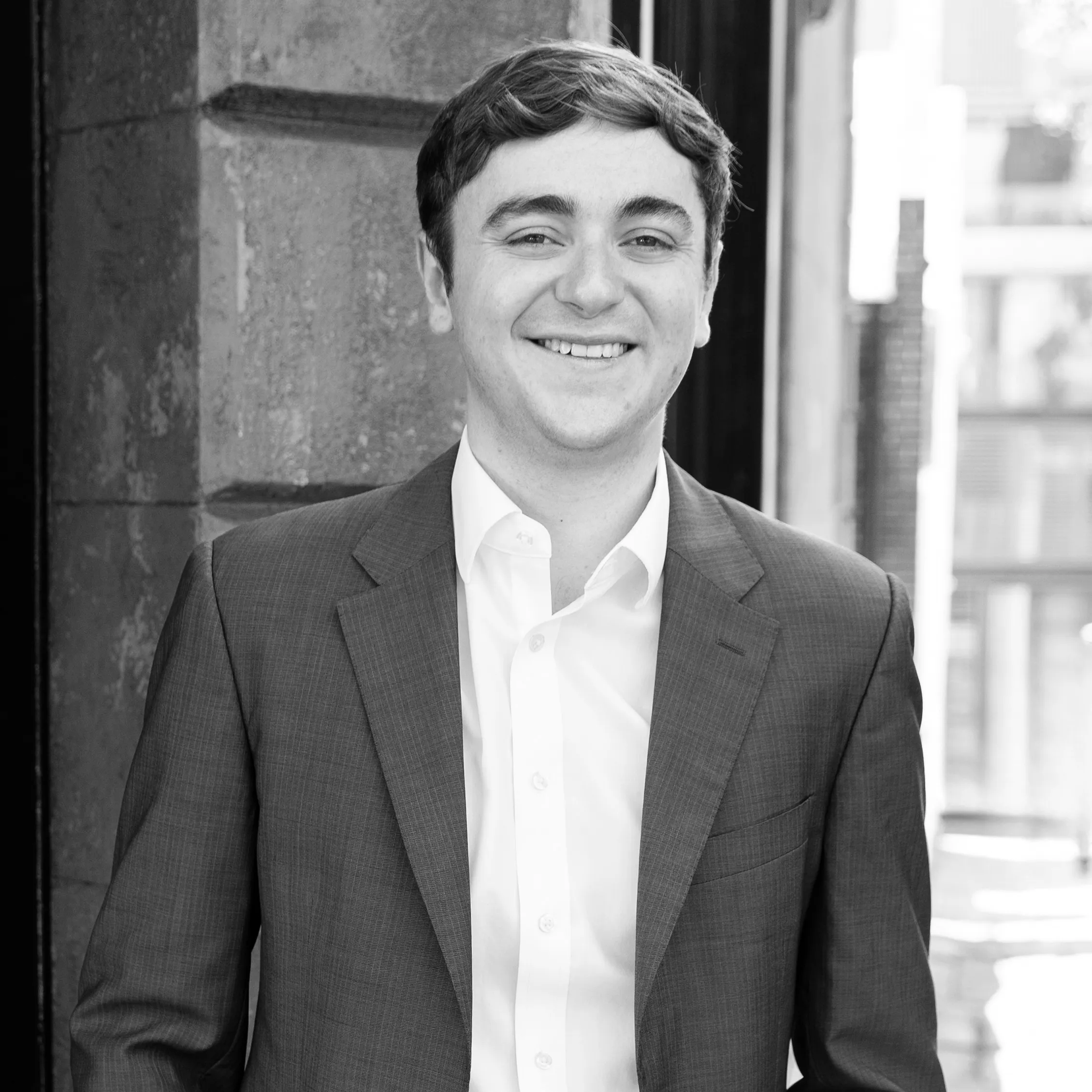 Smiling young man in a suit jacket and white shirt standing against an urban stone wall background.
