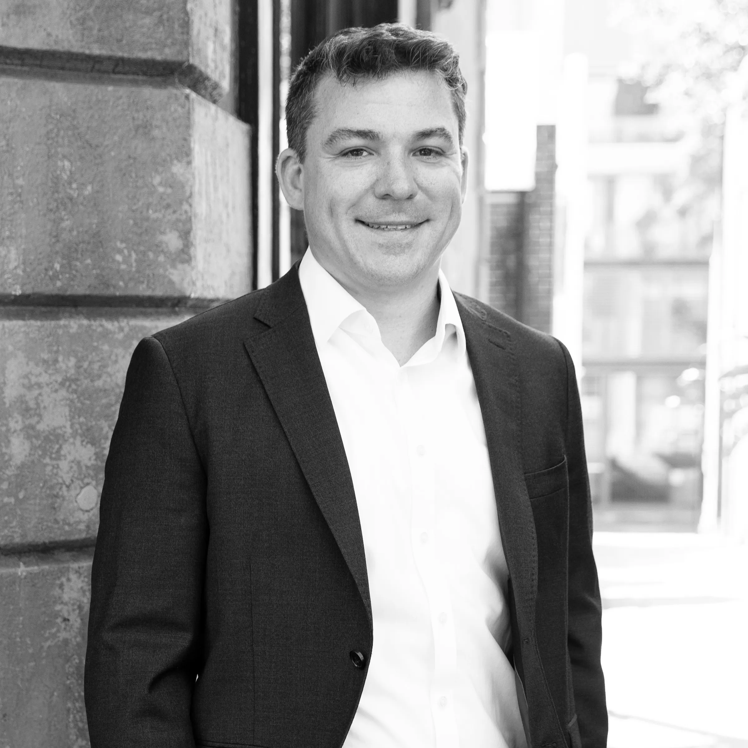 Smiling man wearing a dark suit jacket and white shirt standing outdoors near a building wall.