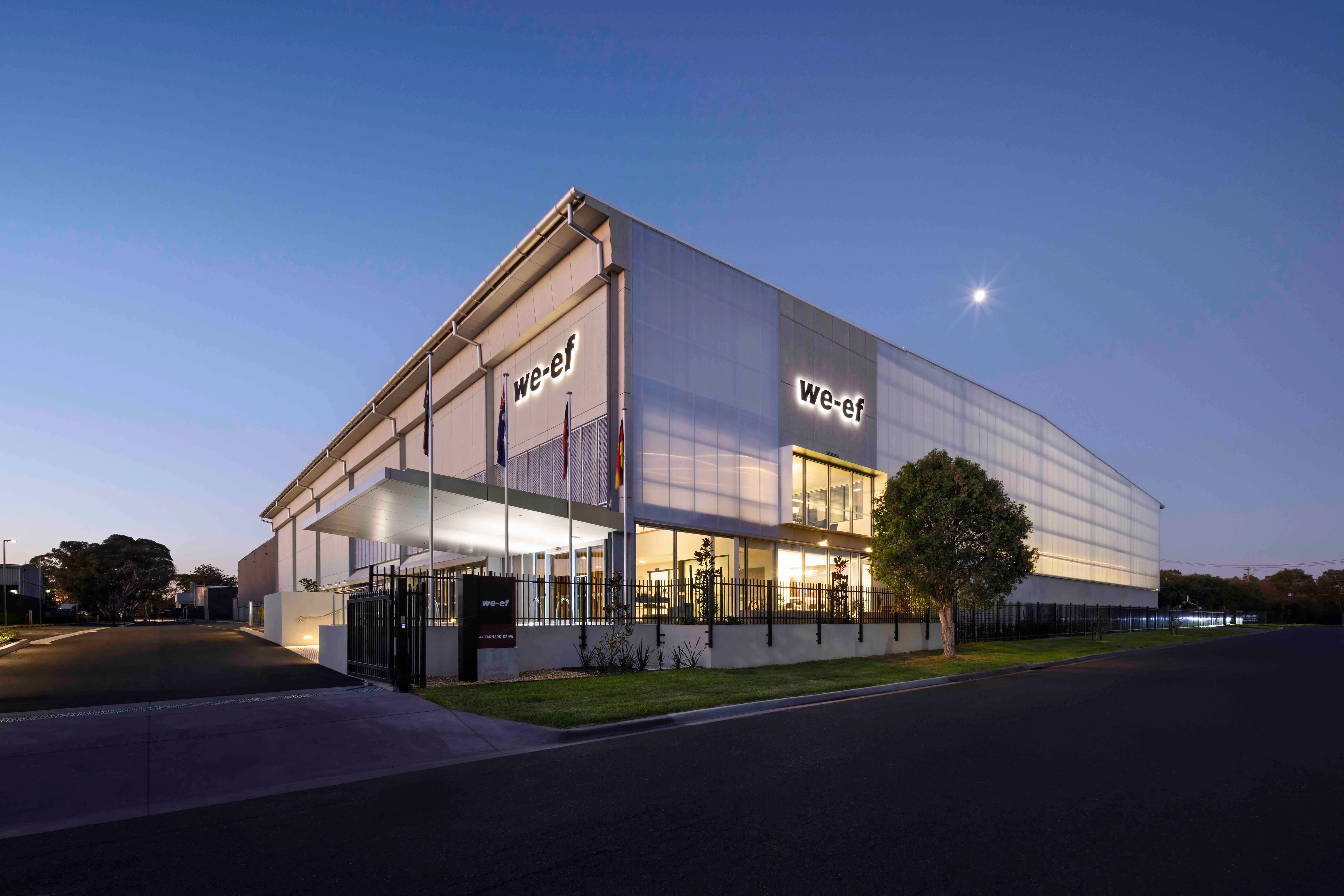 Exterior view of a modern industrial building with illuminated 'we-ef' signs at dusk, with flags and a tree near the entrance.
