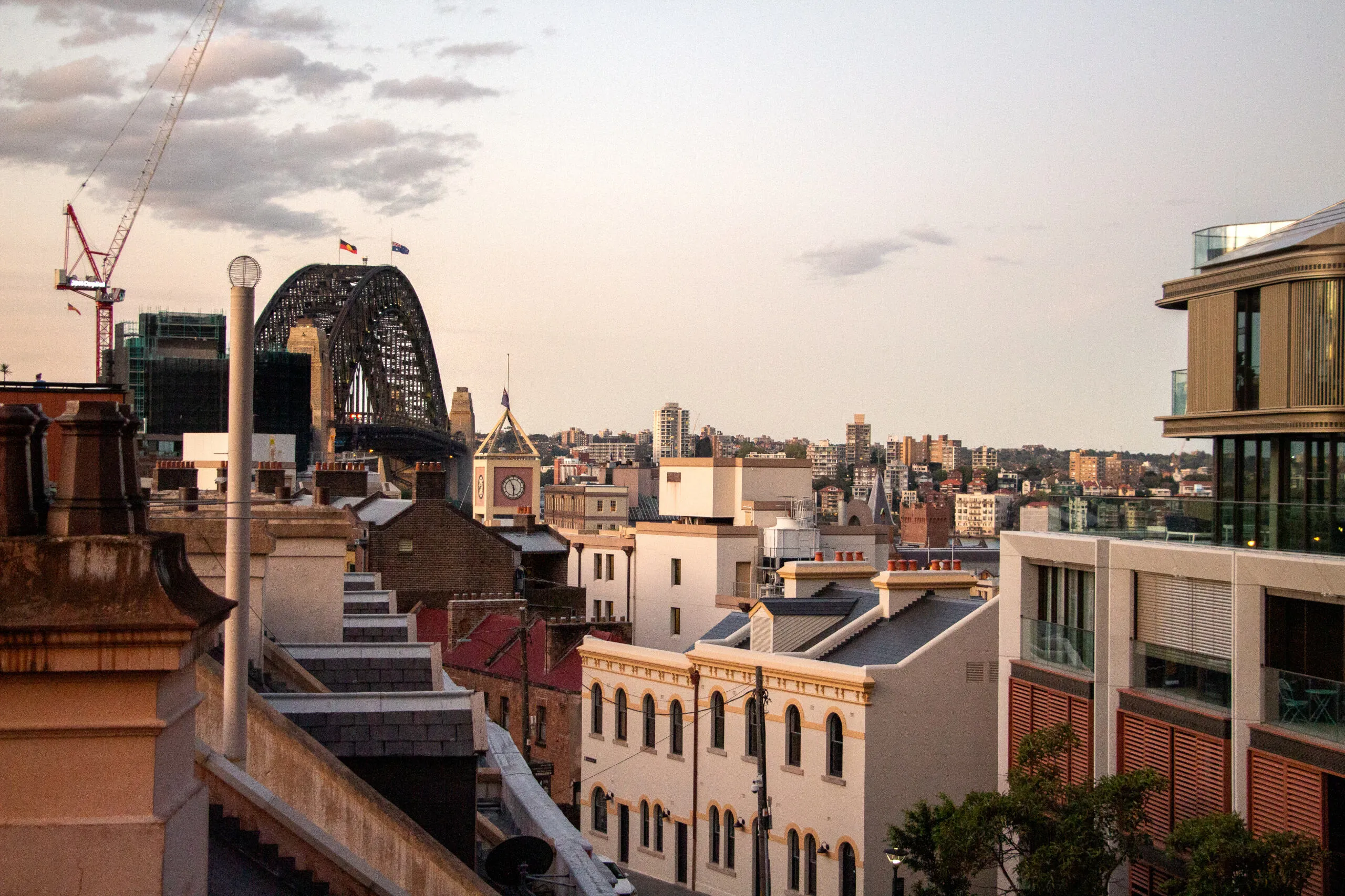 View of Sydney city rooftops at dusk with the Sydney Harbour Bridge and a construction crane in the background.