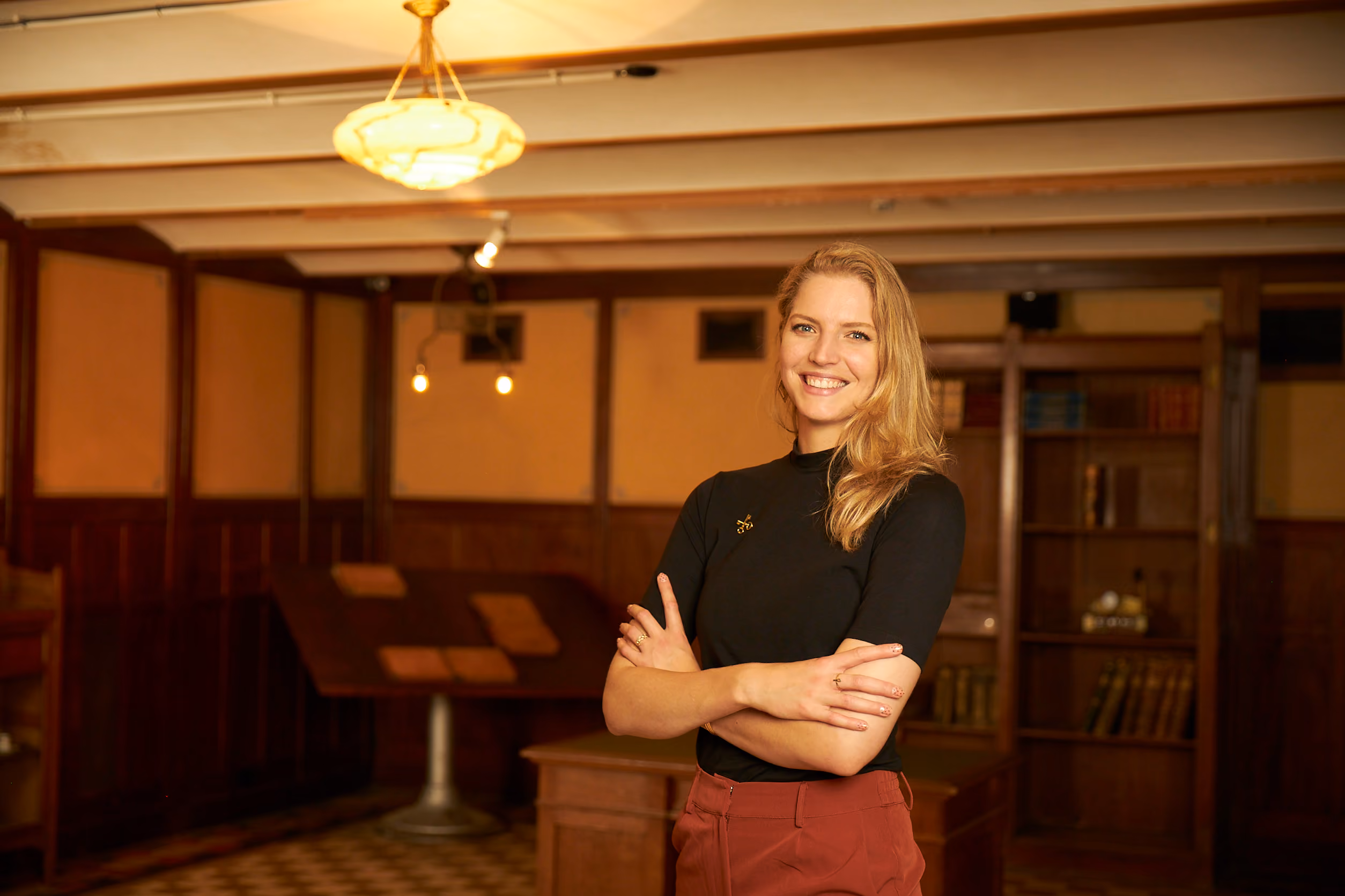 Smiling woman with crossed arms in a warmly lit room with wooden shelves and books.