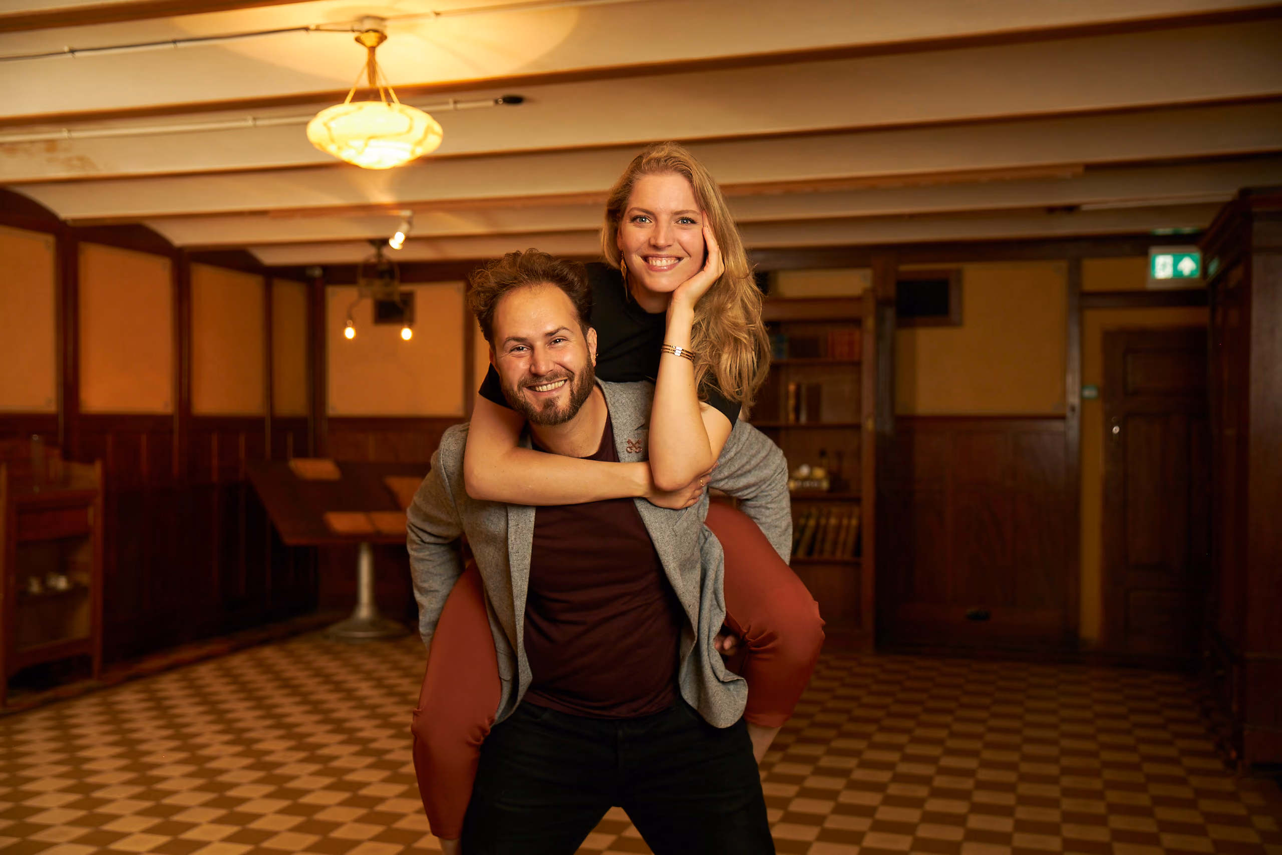 Smiling man giving piggyback ride to smiling woman in a warmly lit room with checkered floor.