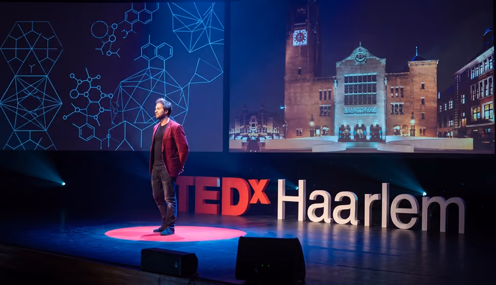 Man in a red jacket stands on stage at TEDx Haarlem with scientific and architectural projections behind him.