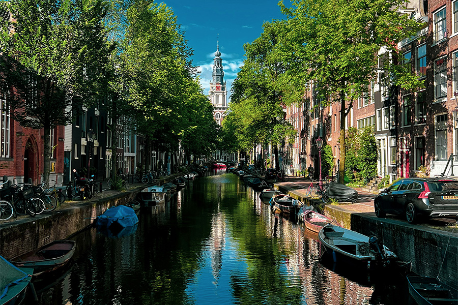 Amsterdam canal view leading to a tower, with boats, vintage houses and trees on both sides of the canal,