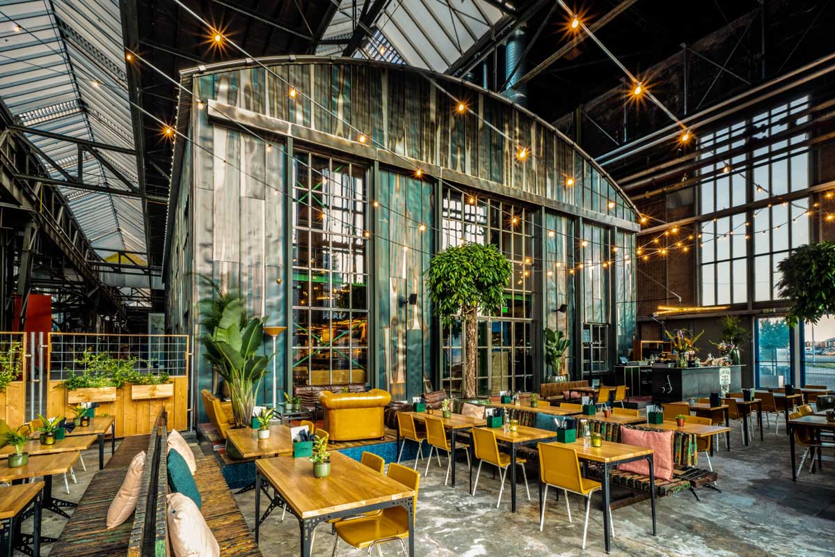 picture of an industrial style restaurant, with tables surrounded by plants and little lights hanging from the ceiling