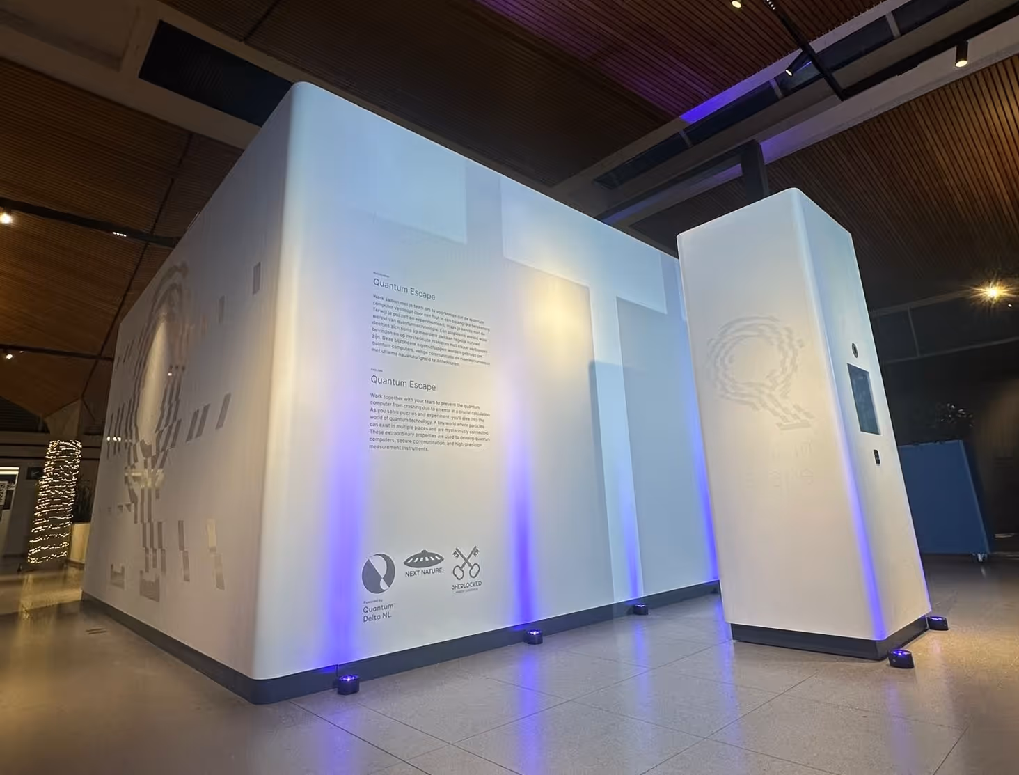 A large, minimalist white cube structure housing the Quantum Escape room, illuminated with blue uplighting and featuring introductory text on its exterior walls.