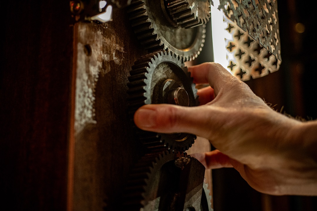A close-up of a player's hand manipulating large, weathered metal gears on a wooden surface, highlighting the tactile and mechanical nature of Sherlocked's puzzle design.