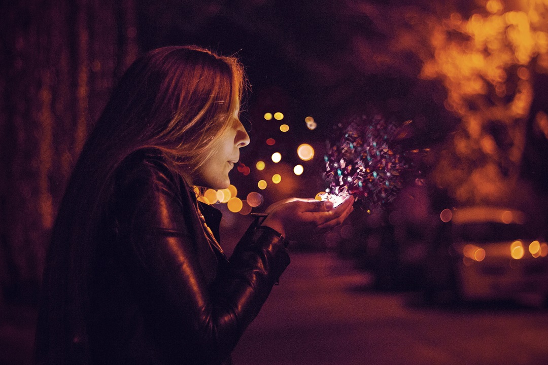 A woman blowing glowing magical sparks from her hands at night, symbolizing Sherlocked’s mission of re-enchanting the world through wonder and play.