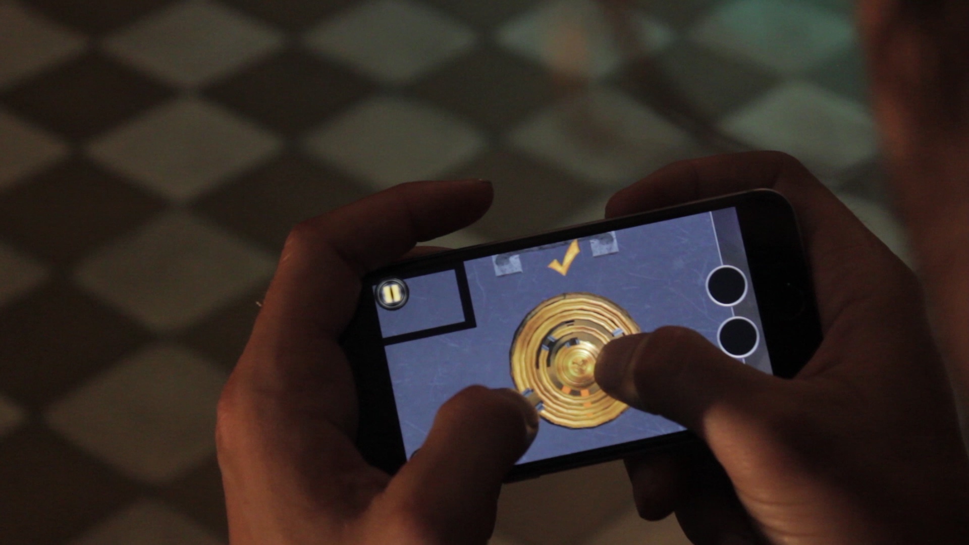 A first-person perspective of a person playing 'The Vault' mobile game on a smartphone. The player's thumbs are interacting with a complex, circular golden ring-lock puzzle on the screen. The background is a blurred, dark room with a checkered floor, emphasizing the immersive atmosphere of the mobile training app designed for escape room preparation.