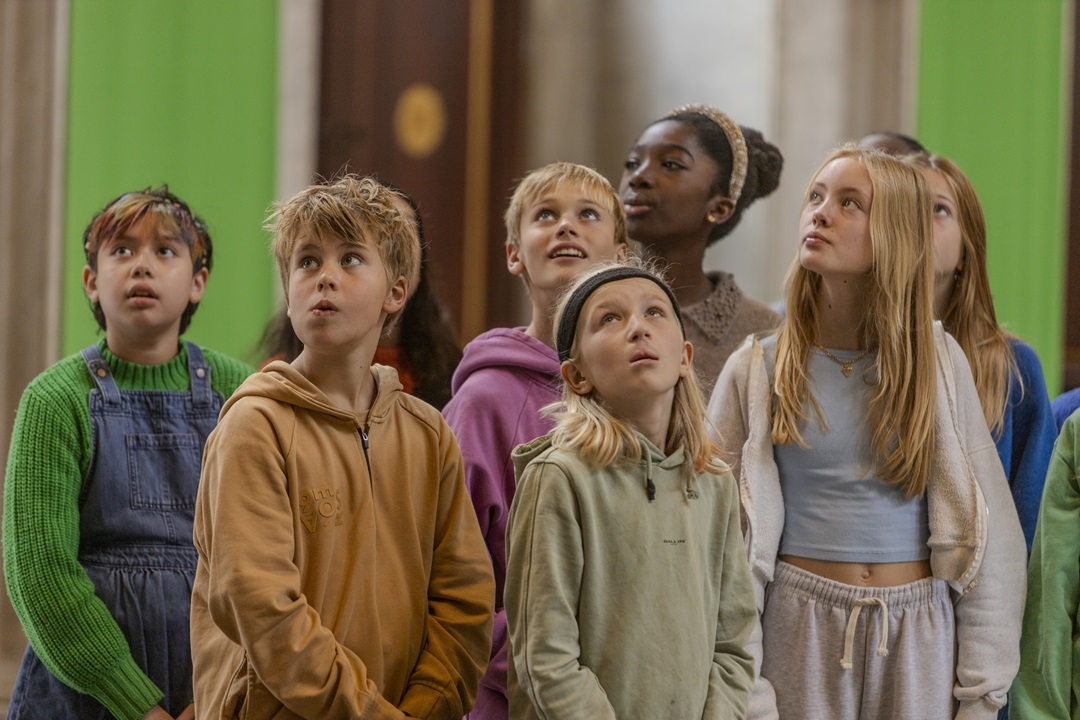 A group of children looking up in wonder at the grand architecture and marble sculptures inside the Citizens' Hall of the Royal Palace Amsterdam during the Palace Mysteries quest.