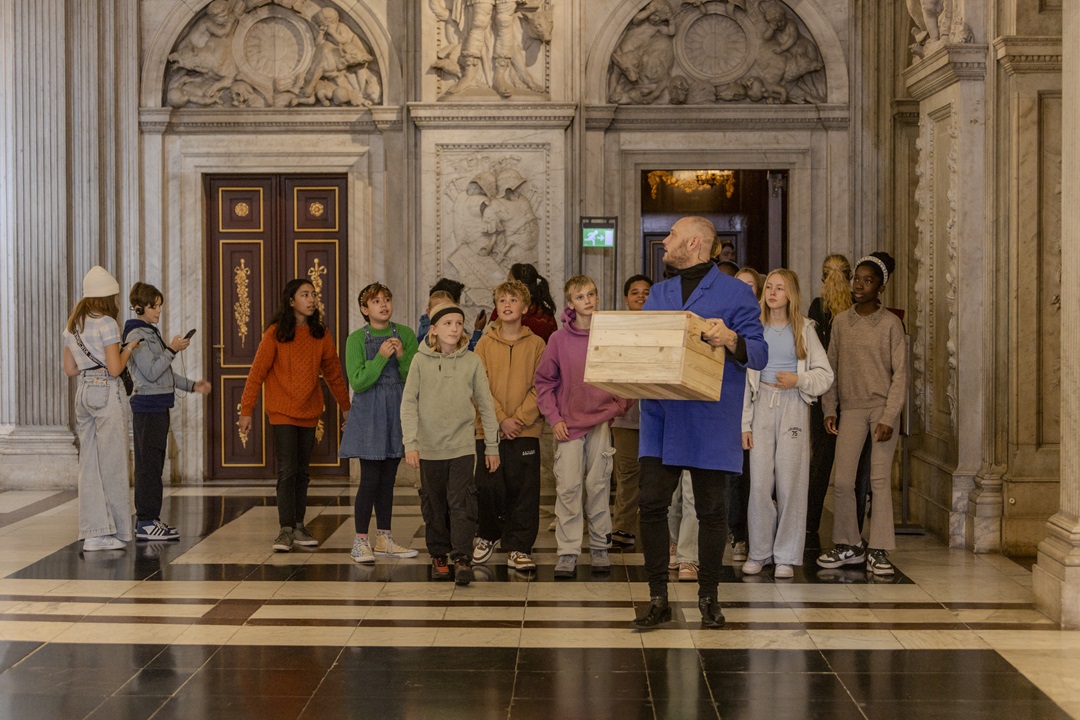 A game master in a blue coat leading a group of school children through the Royal Palace Amsterdam to solve historical puzzles.