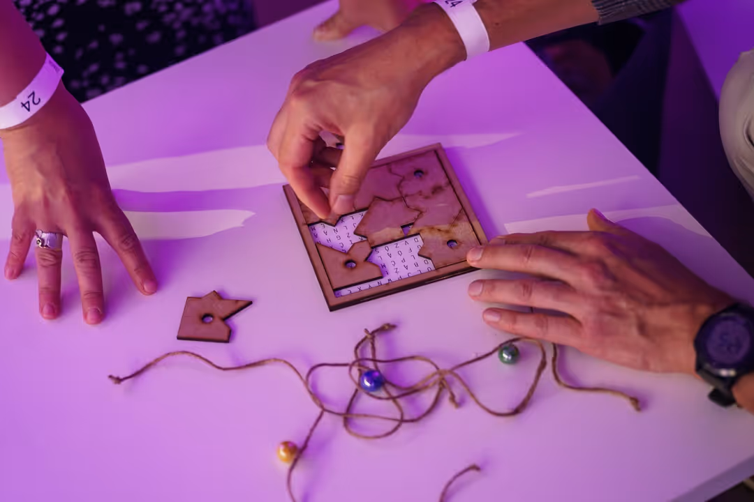 A close-up of hands manipulating a laser-cut wooden jigsaw puzzle on a table. The puzzle pieces are being fitted over a grid of letters as part of a collaborative game design test.