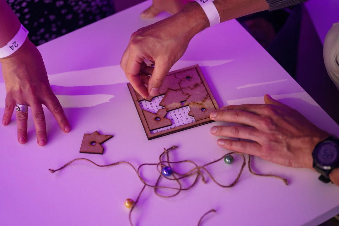 A close-up of hands manipulating a laser-cut wooden jigsaw puzzle on a table. The puzzle pieces are being fitted over a grid of letters as part of a collaborative game design test.