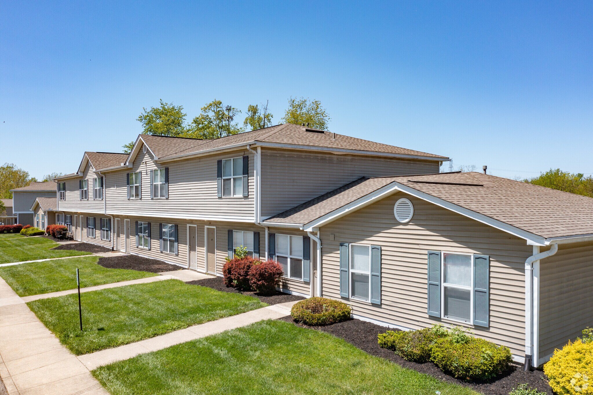 Two-story beige apartment building with blue shutters surrounded by green lawn and shrubs under clear sky.