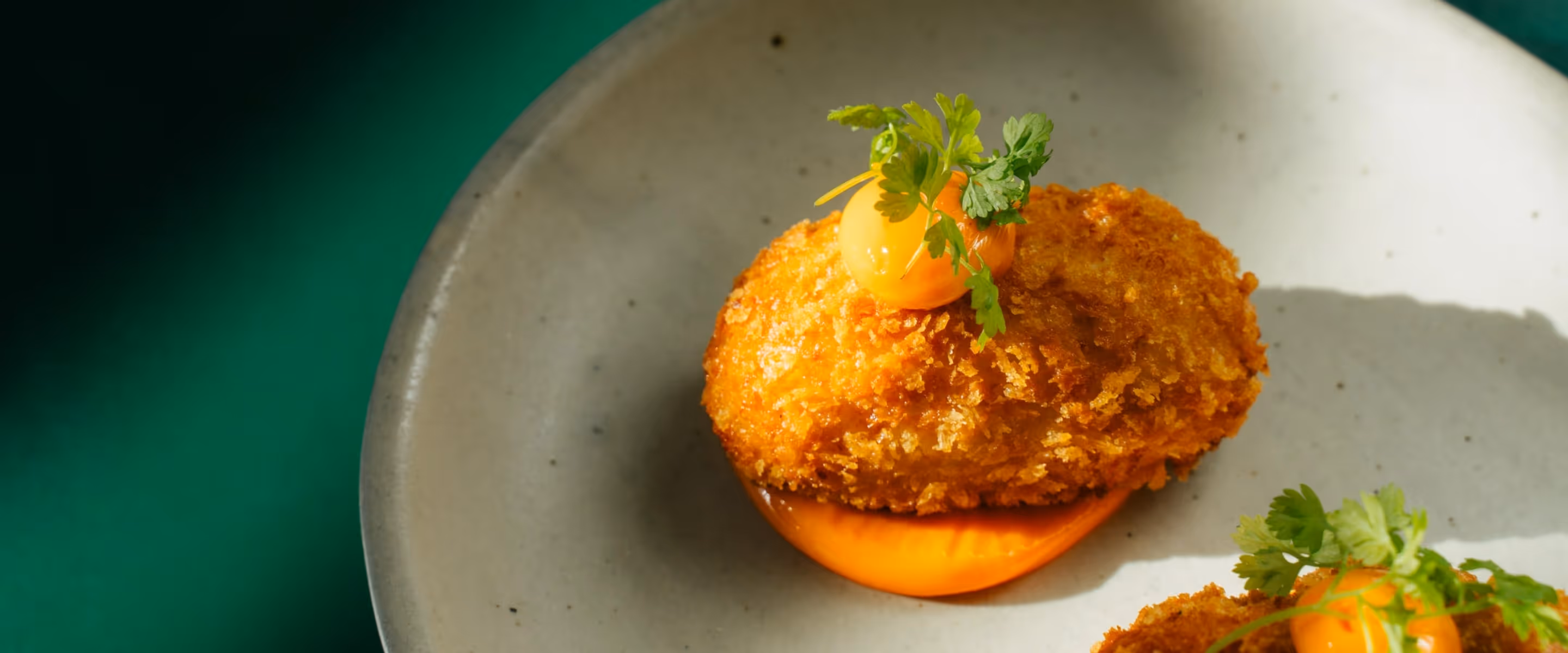 Close-up of a golden, crispy fried croquette served on a dollop of orange sauce and topped with fresh herbs on a ceramic plate.