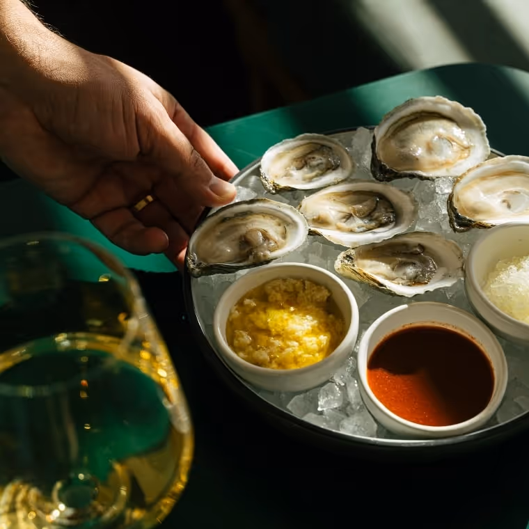 Hand reaching for fresh oysters on the half shell served on ice with mignonette and citrus sauces, beside a glass of white wine on a green table.