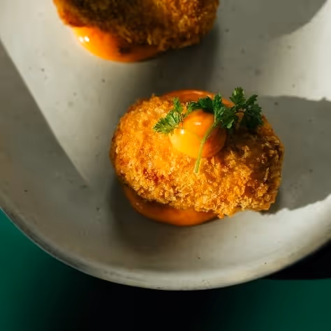 Golden crispy croquette topped with a soft egg yolk and fresh herbs, served with orange sauce on a speckled ceramic plate in natural light.