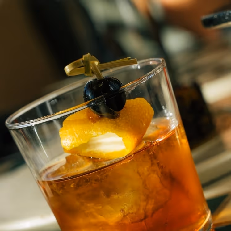 Old fashioned cocktail with ice, orange peel garnish, and a dark cherry on a pick, served in a rocks glass in warm bar light.