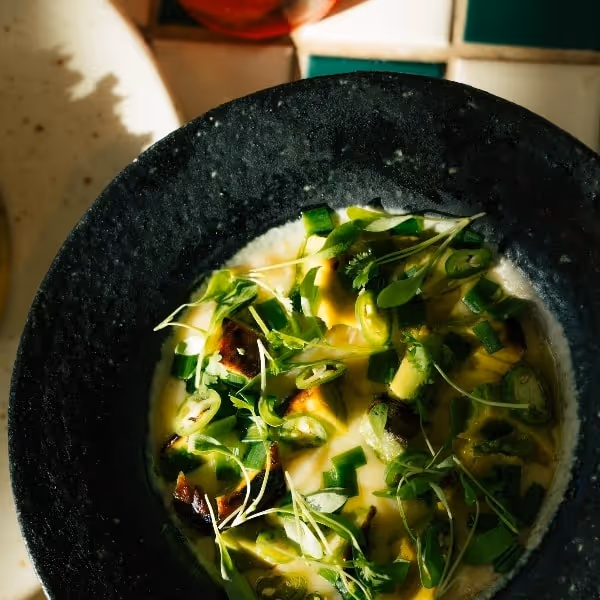 Creamy green soup topped with grilled vegetables, sliced scallions, and fresh herbs, served in a black bowl in warm natural light.