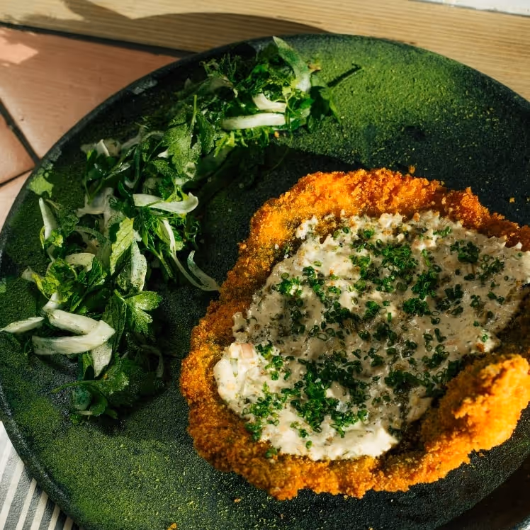 Crispy chicken milanesa topped with creamy herb sauce and chives, served with a fresh green salad on a dark plate in natural light.