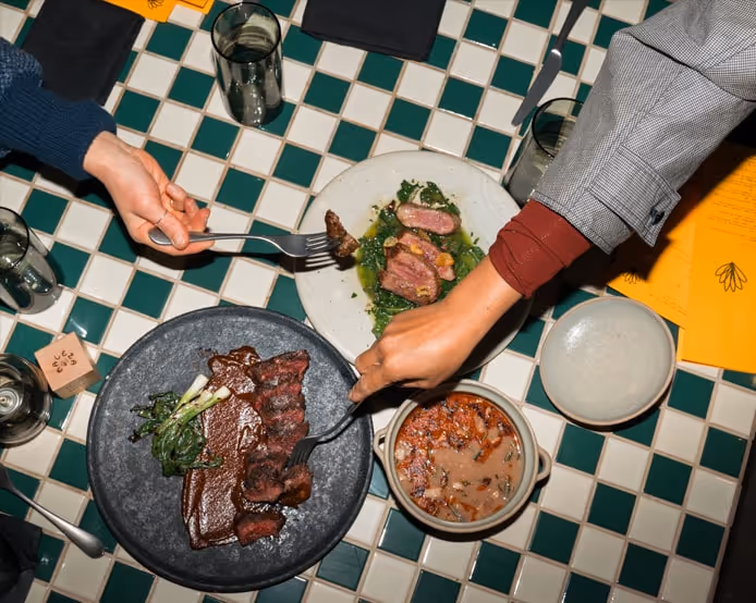 Overhead view of a restaurant dinner table with steaks and sliced meat dishes being served on a green-and-white tiled table.