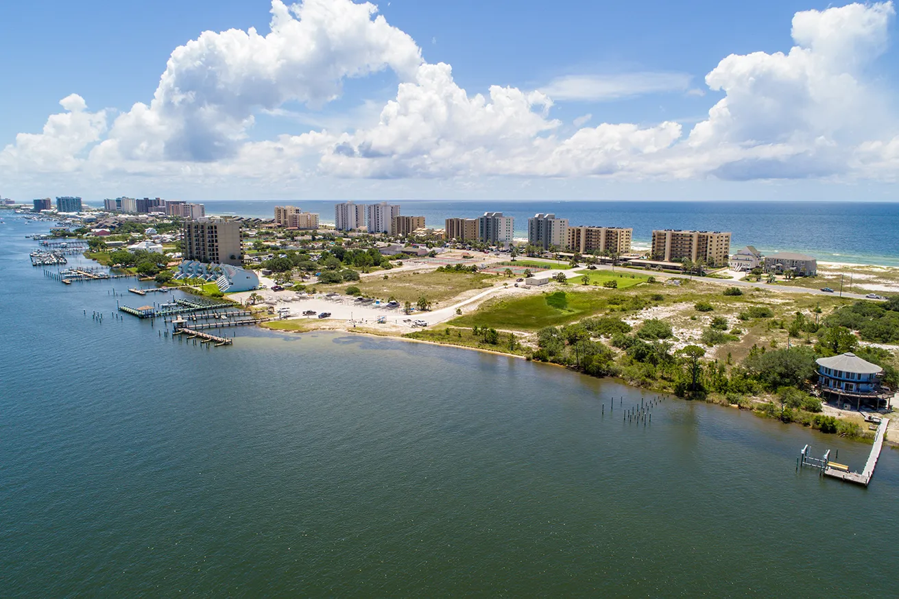 Aerial view of a coastal town with buildings near the shoreline, docks extending into the water, and a partly cloudy sky.