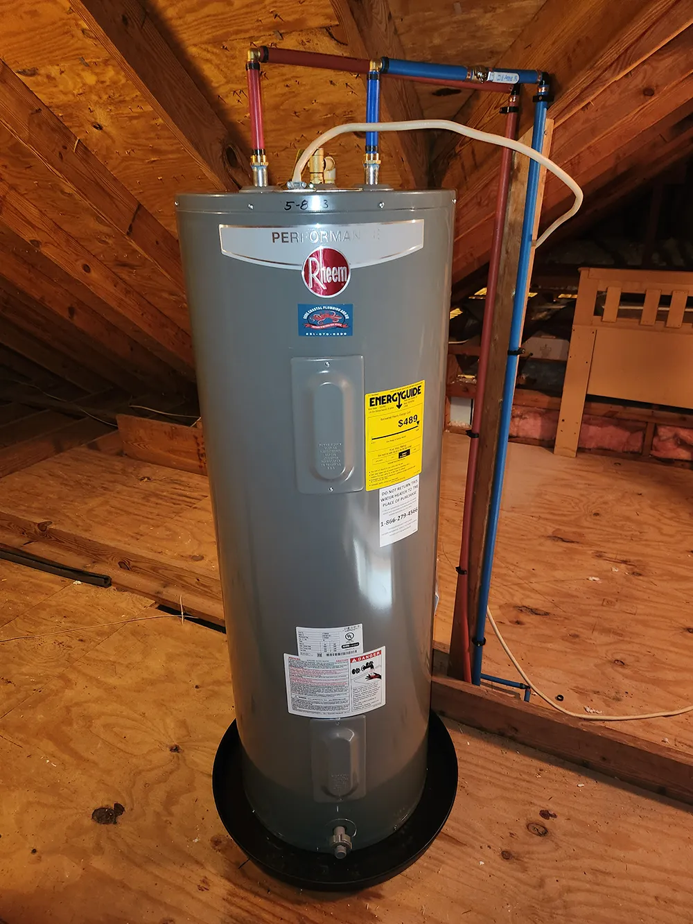 Gray Rheem Performance water heater installed in an attic with red and blue water pipes connected on top.