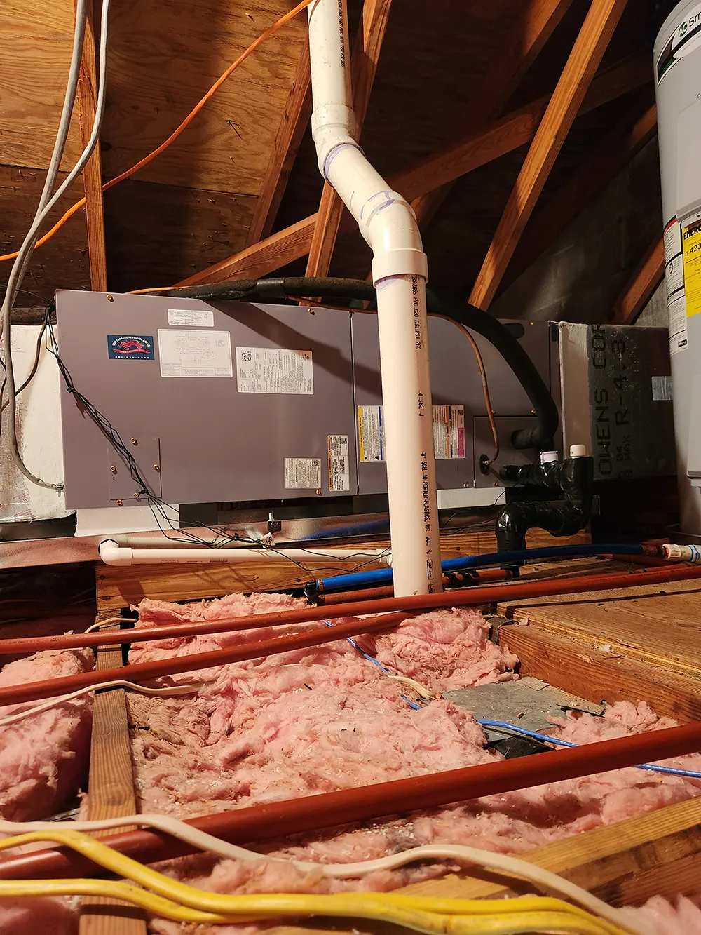 View of attic HVAC unit with PVC pipes, pink insulation, wooden beams, and electrical wiring.