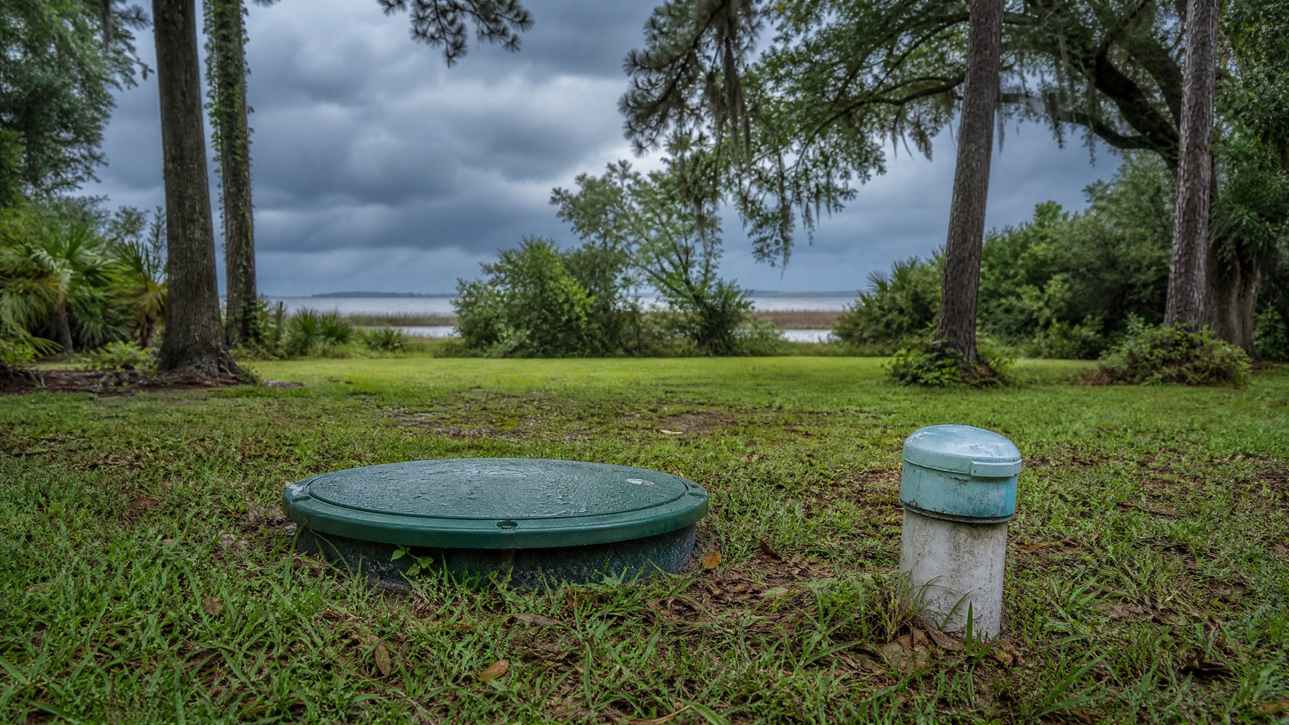 Septic tank access cover and wellhead in a Magnolia Springs yard under approaching storm clouds.