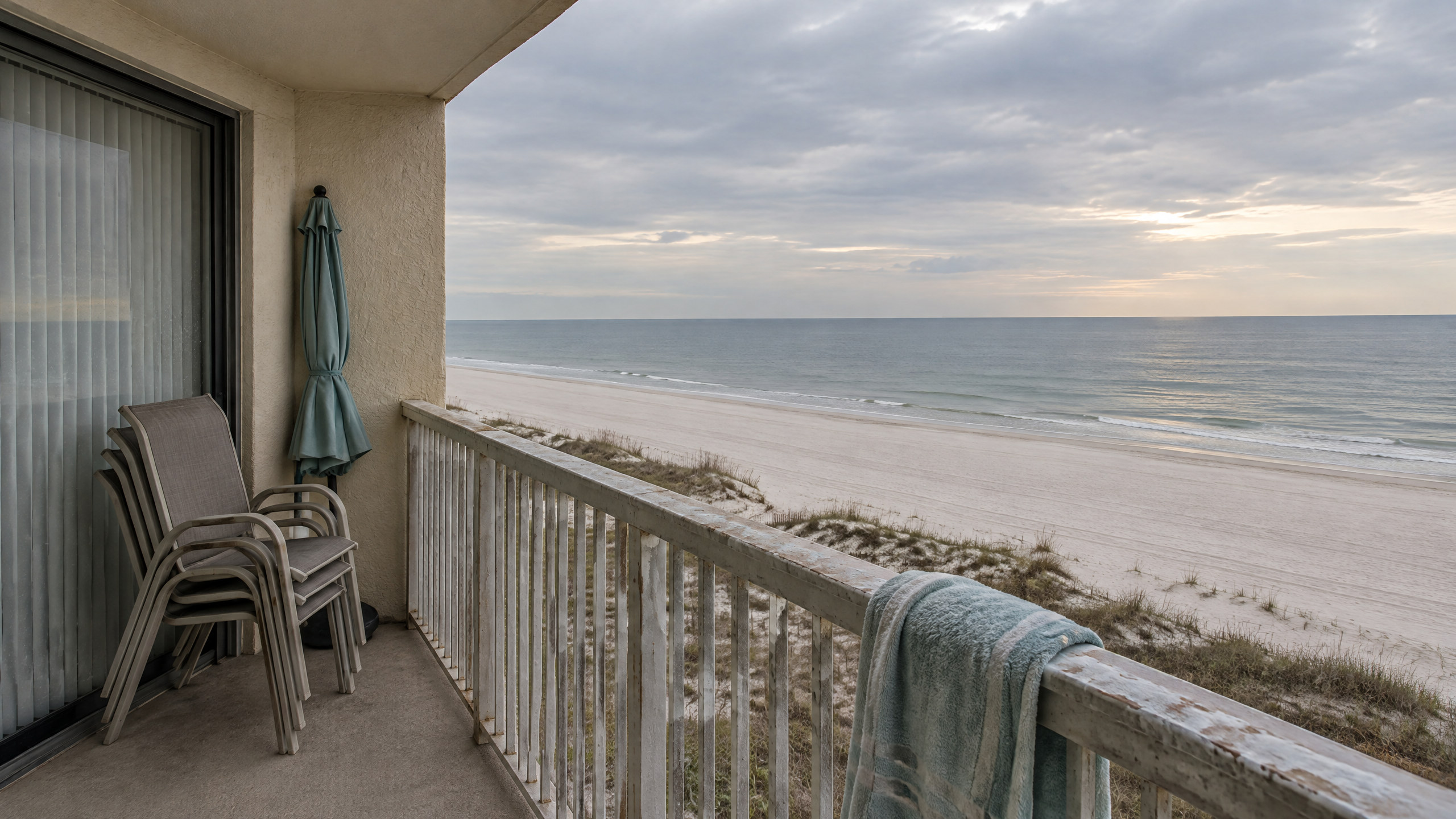 An empty off-season balcony above a quiet Gulf Shores beach, patio chairs stacked and the sliding door shuttered