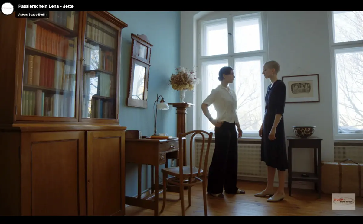 Zwei Frauen stehen sich in einem hellen Raum mit Holzfußboden gegenüber. Links steht ein großer Holzschrank, daneben ein alter Schreibtisch mit Lampe und Stuhl. Rechts sorgen große Fenster für natürliches Licht.