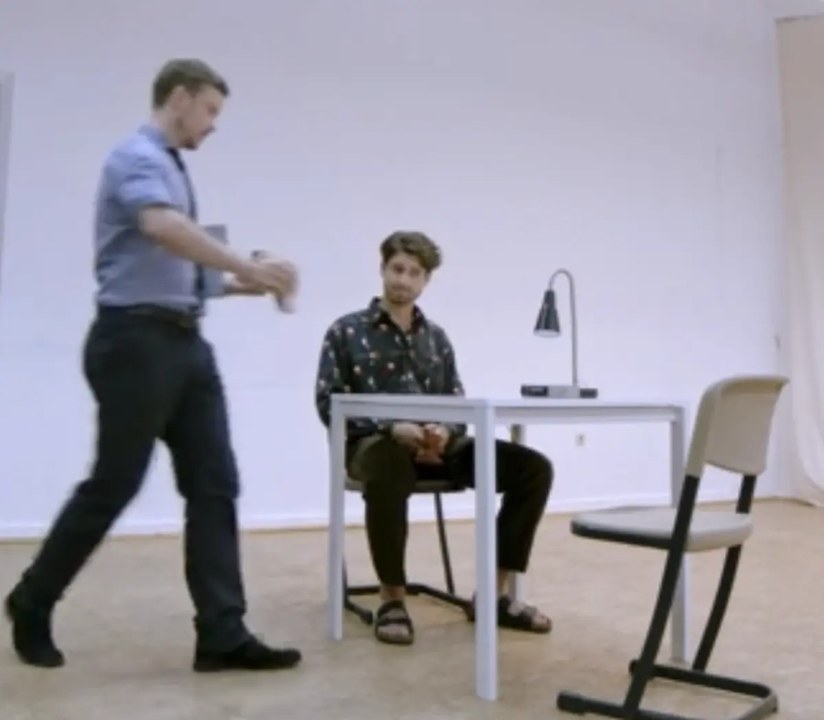 Ein Mann sitzt an einem Tisch in einem leeren, hellen Raum. Ein anderer Mann geht im Vordergrund vorbei. Neben dem Tisch steht ein einzelner Stuhl und eine kleine Schreibtischlampe.