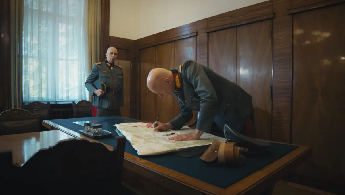 Zwei Männer in historischen Militäruniformen befinden sich in einem holzvertäfelten Raum. Einer steht im Hintergrund, der andere beugt sich über eine große Karte auf einem Tisch und schreibt darauf.