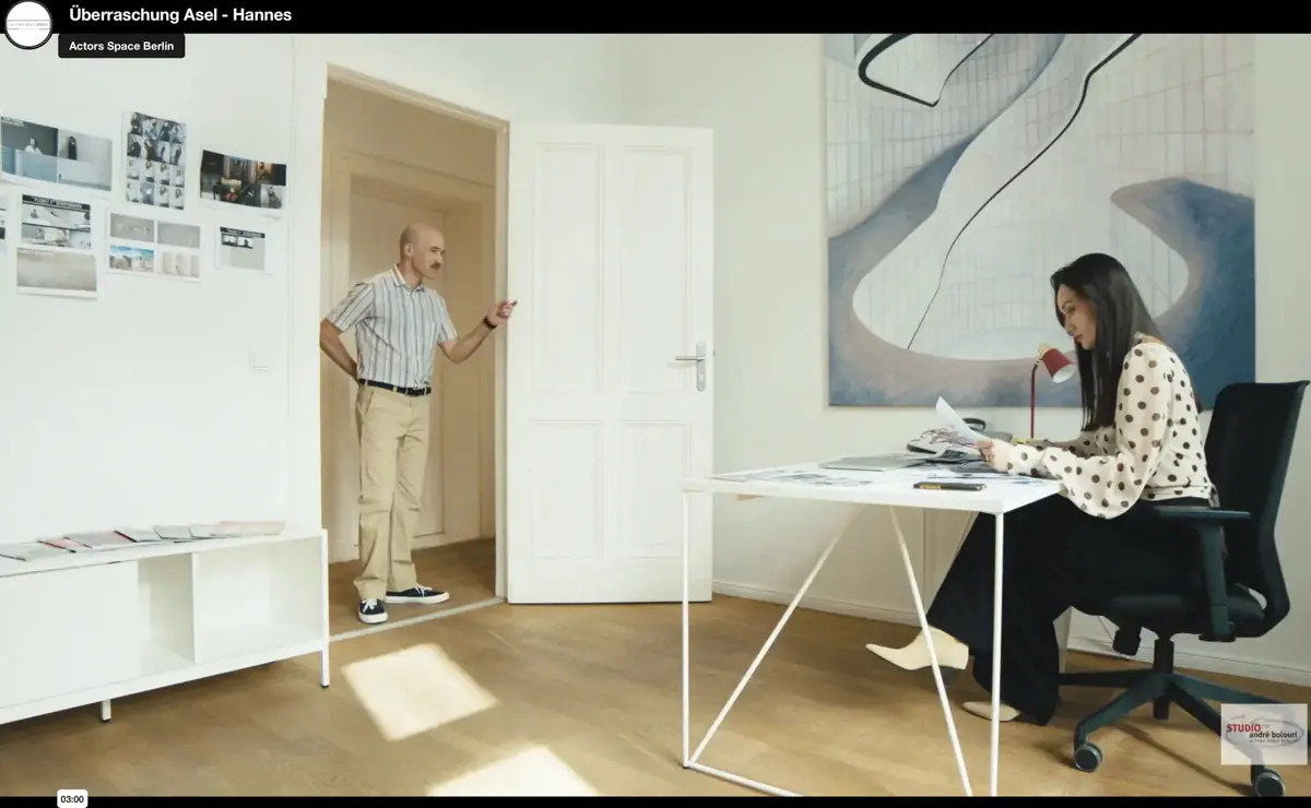 In einem hellen Büro sitzt eine Frau an einem modernen Schreibtisch und sortiert Unterlagen, während ein Mann im Türrahmen steht und sie anspricht. An der Wand hängen Moodboards und ein großes abstraktes Bild.