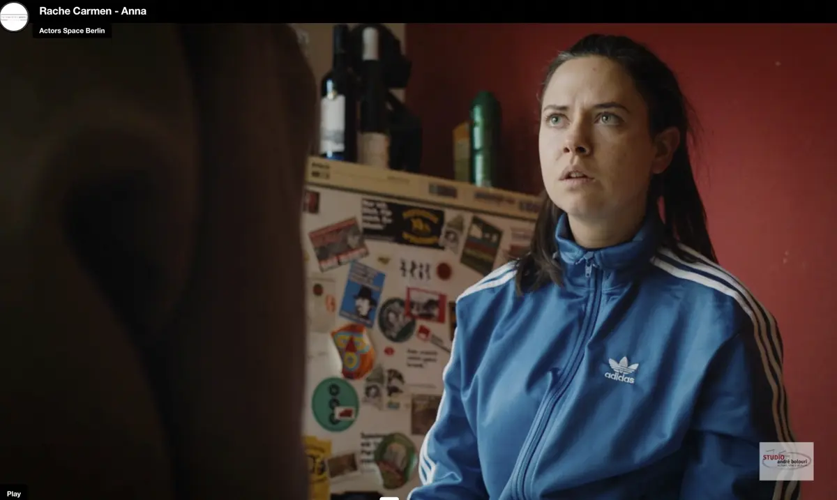 Eine Frau in einer blauen Trainingsjacke steht in einer Küche vor einem Kühlschrank mit vielen Stickern und Blickt überrascht oder irritiert nach oben. Im Vordergrund ist die Schulter einer zweiten Person unscharf zu sehen.