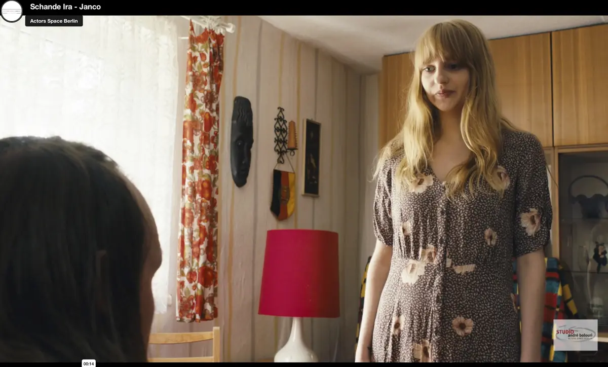 Eine blonde Frau in einem grünen Shirt steht in einem grauen, backsteinartigen Raum und spricht mit einer anderen Person, die nur von hinten zu sehen ist.