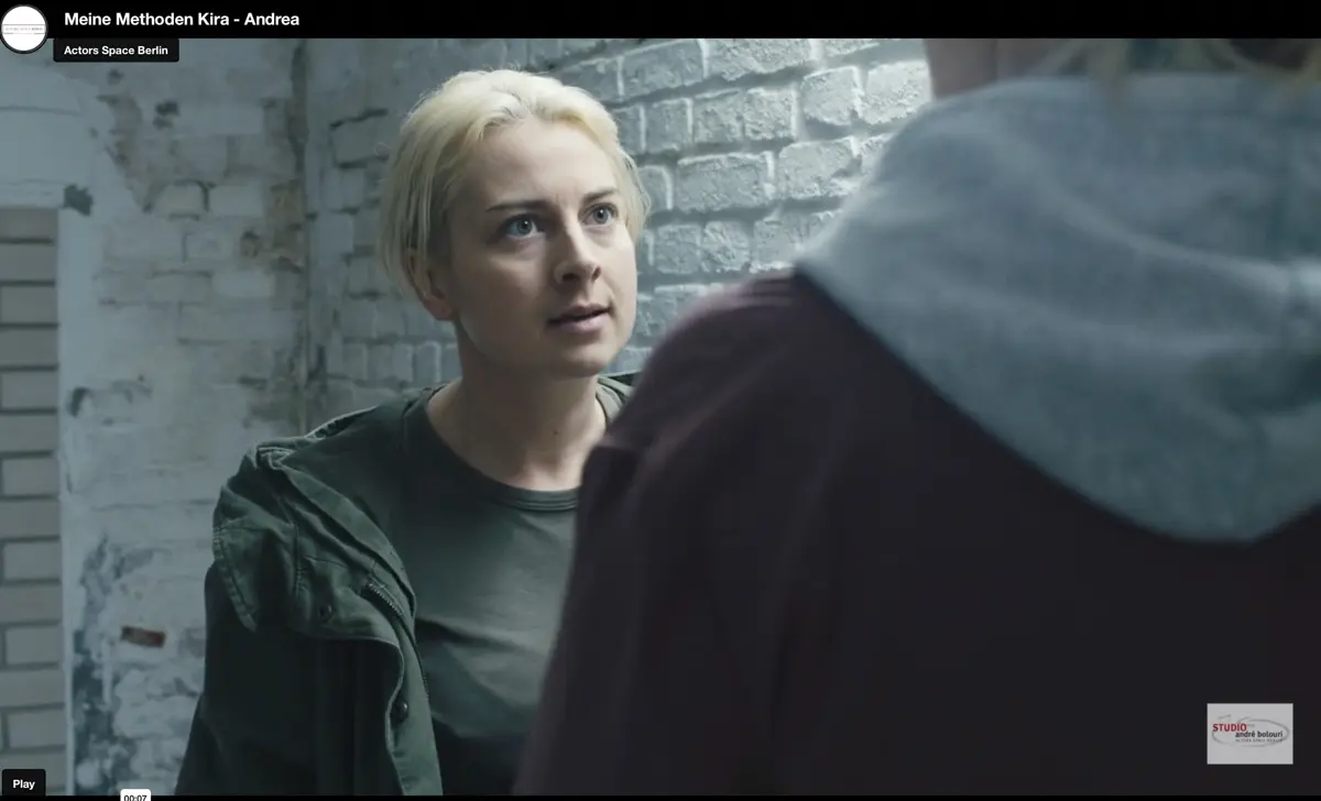 Eine junge Frau mit langen blonden Haaren steht in einem gemütlichen, nostalgisch eingerichteten Zimmer. Rechts stehen Holzschränke, links ein Fenster mit gemustertem Vorhang und eine rote Tischlampe.