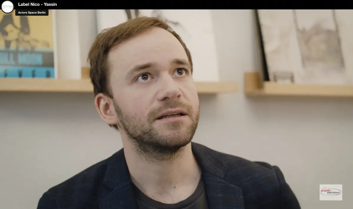 Nahaufnahme eines Mannes, der nach oben schaut. Er sitzt in einem hellen Raum mit minimalistischer Einrichtung, im Hintergrund hängen Bücher und Bilderrahmen auf schmalen Holzleisten.