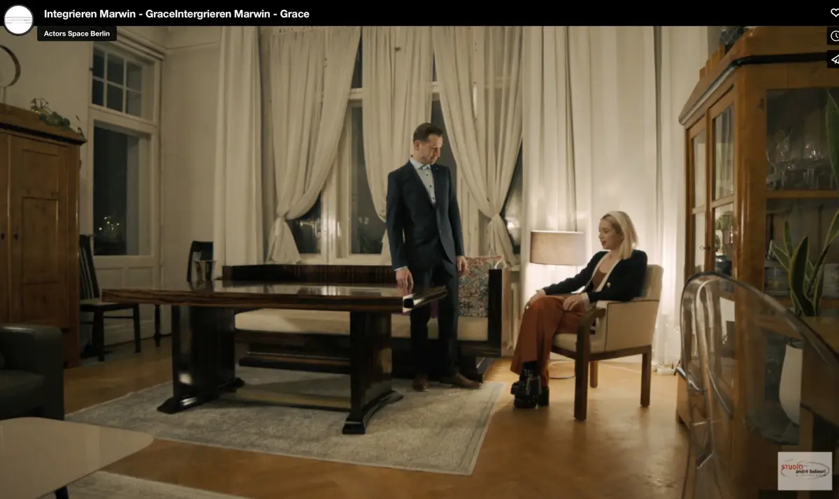 Ein Mann im Anzug steht an einem großen dunklen Holztisch in einem eleganten Wohnzimmer mit hohen Fenstern und weißen Vorhängen. Eine Frau sitzt ihm gegenüber auf einem Polsterstuhl und wirkt nachdenklich. Die Szene ist warm beleuchtet.